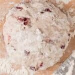 Ball of floured bread dough with cranberries on a wooden surface, surrounded by scattered flour and a metal bench scraper.