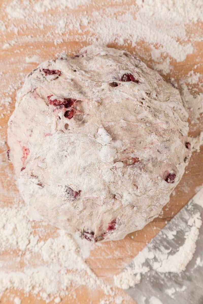 Ball of floured bread dough with cranberries on a wooden surface, surrounded by scattered flour and a metal bench scraper.