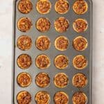 A baking tray filled with 24 mini pecan tassies, arranged in a 4 by 6 grid, on a light countertop.