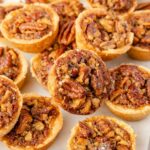 Mini pecan tarts arranged on a white surface, each filled with a baked pecan and syrup mixture, with some whole pecans scattered around them.
