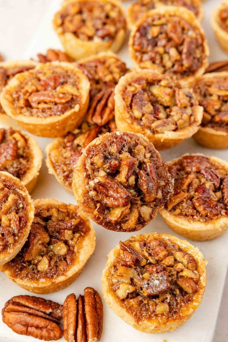 Mini pecan tarts arranged on a white surface, each filled with a baked pecan and syrup mixture, with some whole pecans scattered around them.