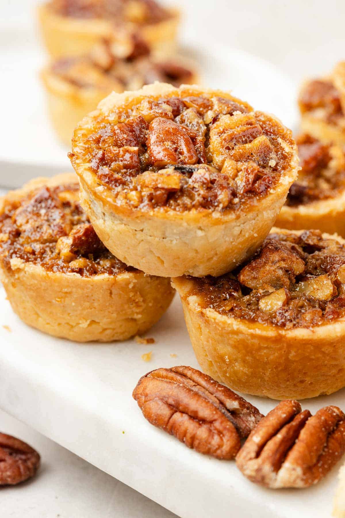 Close-up of mini pecan tassies stacked on a white surface, with whole pecans scattered nearby.