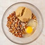 A glass bowl containing chopped pecans, brown sugar, and a raw egg on a light-colored surface.