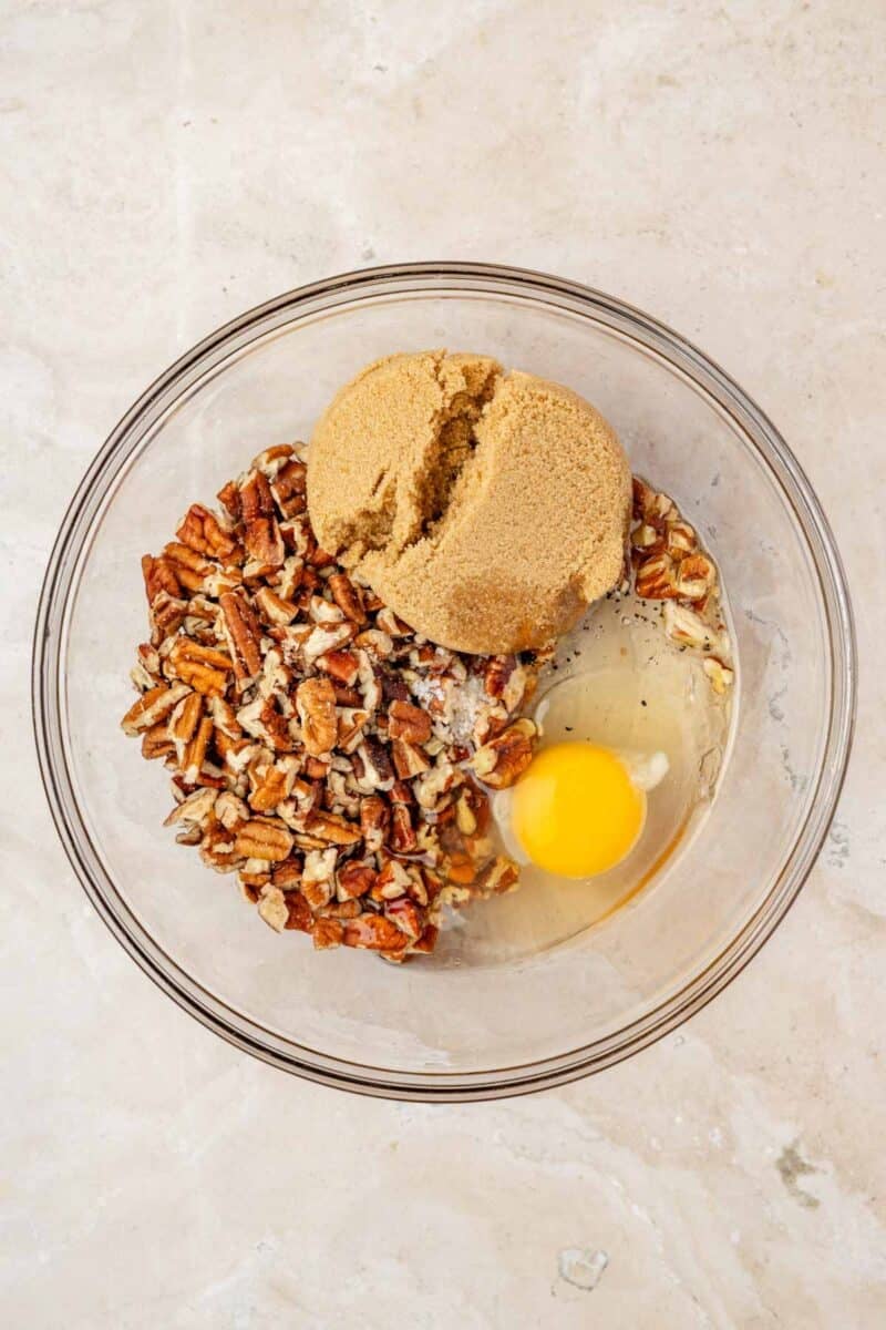 A glass bowl containing chopped pecans, brown sugar, and a raw egg on a light-colored surface.