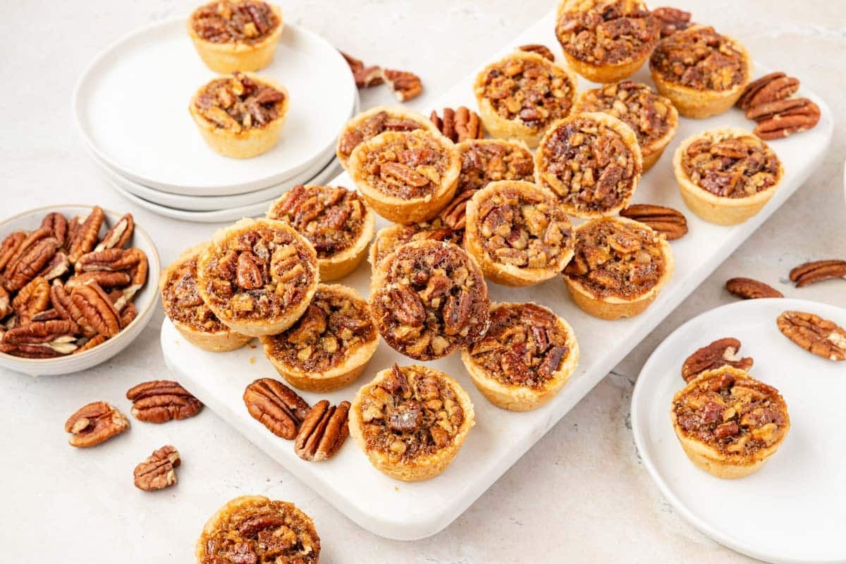 Mini pecan tassies are arranged on a white board with several pecans scattered around, surrounded by white plates and small bowls of pecans.