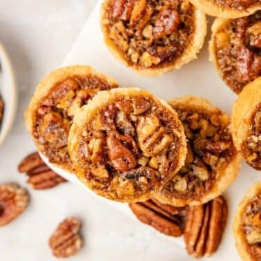 Mini pecan tassies arranged on a white surface, with whole pecans scattered nearby and a bowl of pecans in the background.