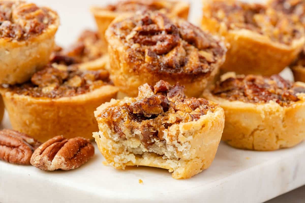 A group of mini pecan pies sits on a white surface, with one in the front showing a bite taken out, revealing the nutty filling and flaky crust.