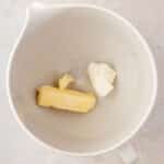 A white mixing bowl containing a stick of butter and a chunk of cream cheese on a light countertop.