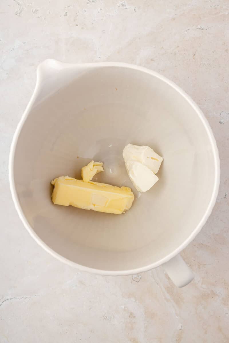 A white mixing bowl containing a stick of butter and a chunk of cream cheese on a light countertop.