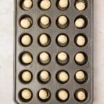 A mini muffin tin with 24 wells, each holding a single round ball of dough, placed on a light-colored surface.