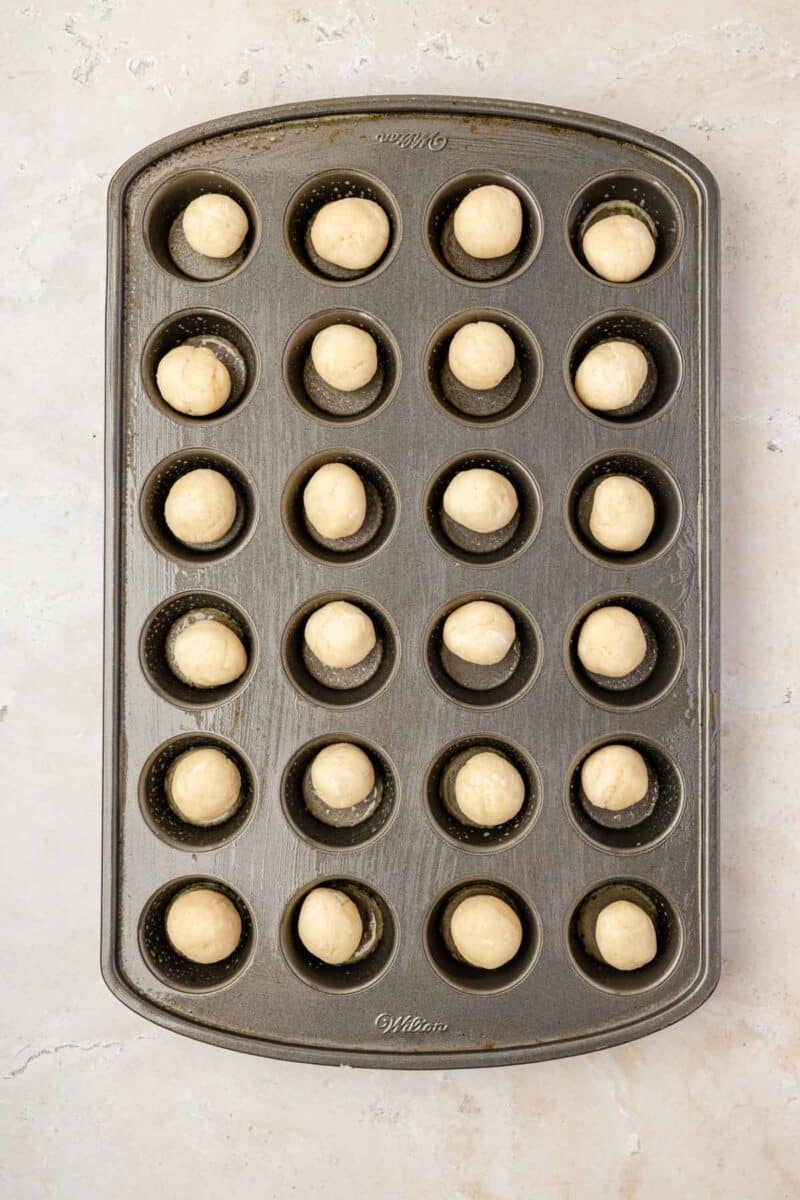 A mini muffin tin with 24 wells, each holding a single round ball of dough, placed on a light-colored surface.