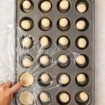 A hand lifts plastic wrap covering a muffin tin filled with round balls of dough, arranged in each cup on a light surface.