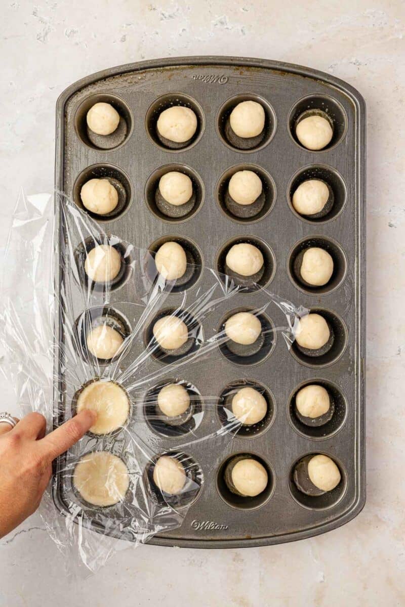 A hand lifts plastic wrap covering a muffin tin filled with round balls of dough, arranged in each cup on a light surface.