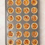 A muffin tin filled with unbaked mini pecan tassies, each cup containing pie crust and pecan filling, placed on a light-colored surface.