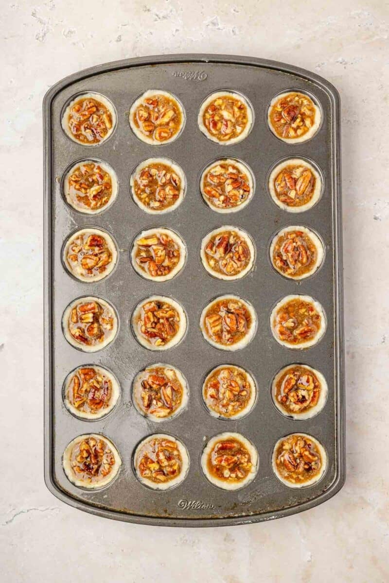 A muffin tin filled with unbaked mini pecan tassies, each cup containing pie crust and pecan filling, placed on a light-colored surface.
