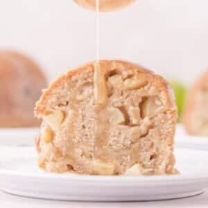 A slice of apple cake on a white plate with glaze being drizzled on top.