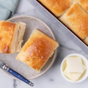 Two buttered dinner rolls on a small plate next to a baking pan of rolls and a dish of butter pats.