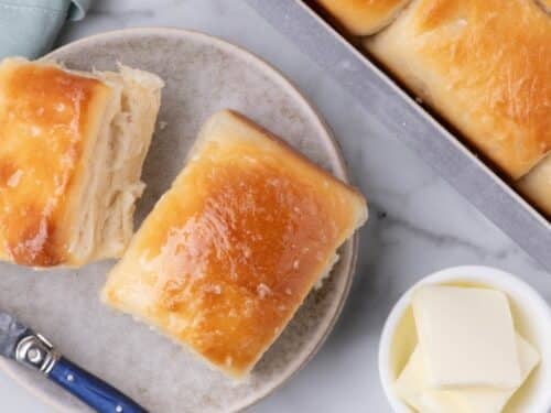 Two buttered dinner rolls on a small plate next to a baking pan of rolls and a dish of butter pats.