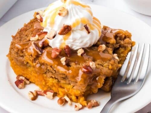 A slice of pumpkin dump cake topped with whipped cream, caramel drizzle, and pecans, served on a white plate with a fork.