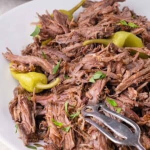 Shredded mississippi pot roast topped with whole pepperoncini peppers and chopped herbs on a white plate.