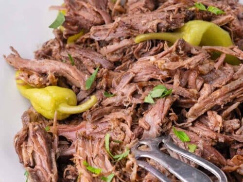 Shredded mississippi pot roast topped with whole pepperoncini peppers and chopped herbs on a white plate.