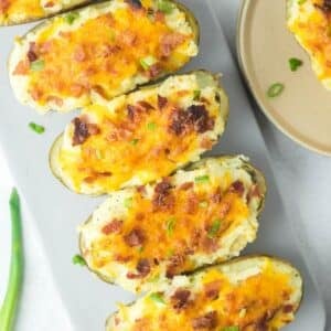 Five twice-baked potatoes topped with melted cheddar cheese, bacon bits, and chopped green onions on a white serving platter.