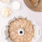 Apple cake batter in a fluted bundt pan, surrounded by bowls containing butter, cream, and sugar.