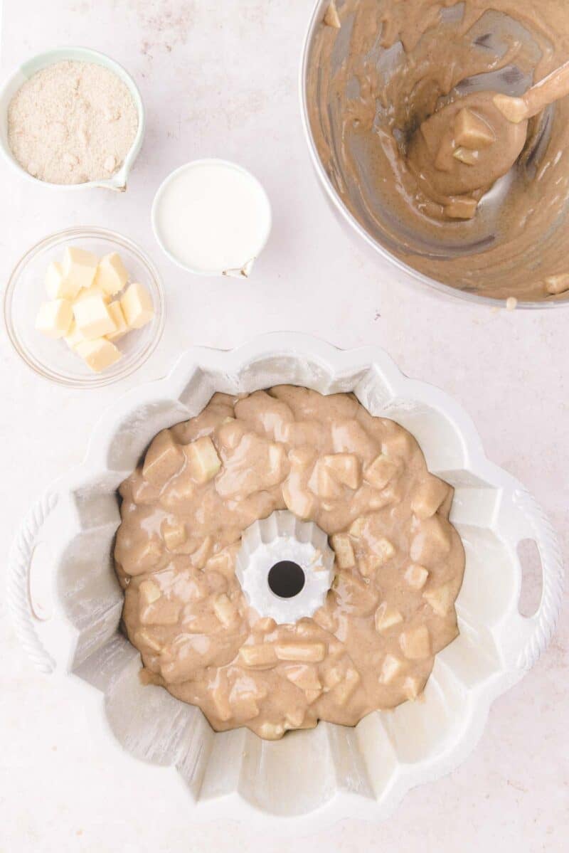 Apple cake batter in a fluted bundt pan, surrounded by bowls containing butter, cream, and sugar.