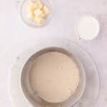 A saucepan filled with sugar sits on a marble board next to a small bowl of cubed butter and a pitcher of milk.