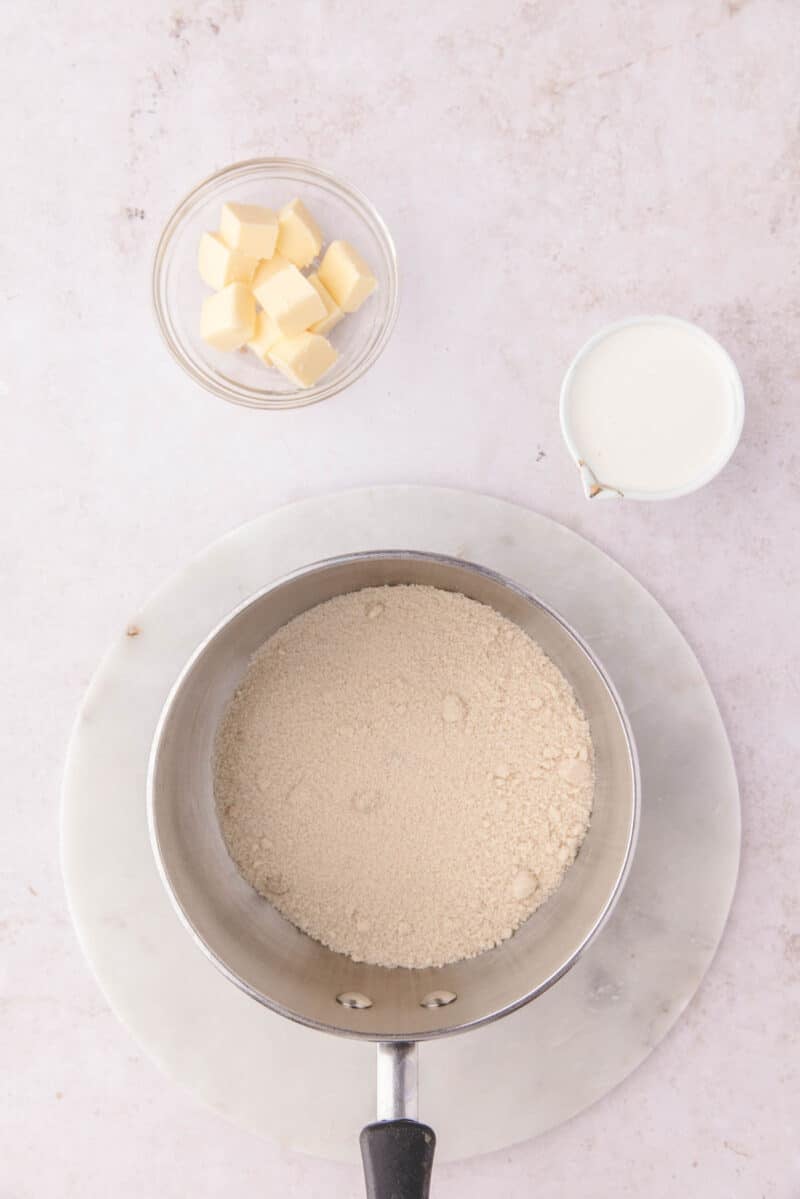 A saucepan filled with sugar sits on a marble board next to a small bowl of cubed butter and a pitcher of milk.