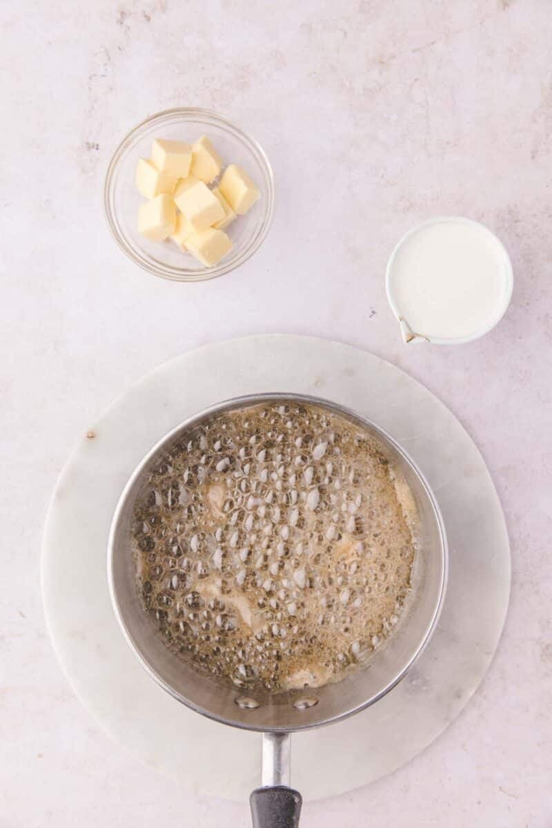 A saucepan of bubbling caramel on a marble board, with a small bowl of butter cubes and a small pitcher of cream nearby.
