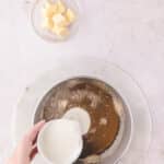 A hand pours cream into a saucepan containing browned butter, with a small bowl of butter cubes nearby.