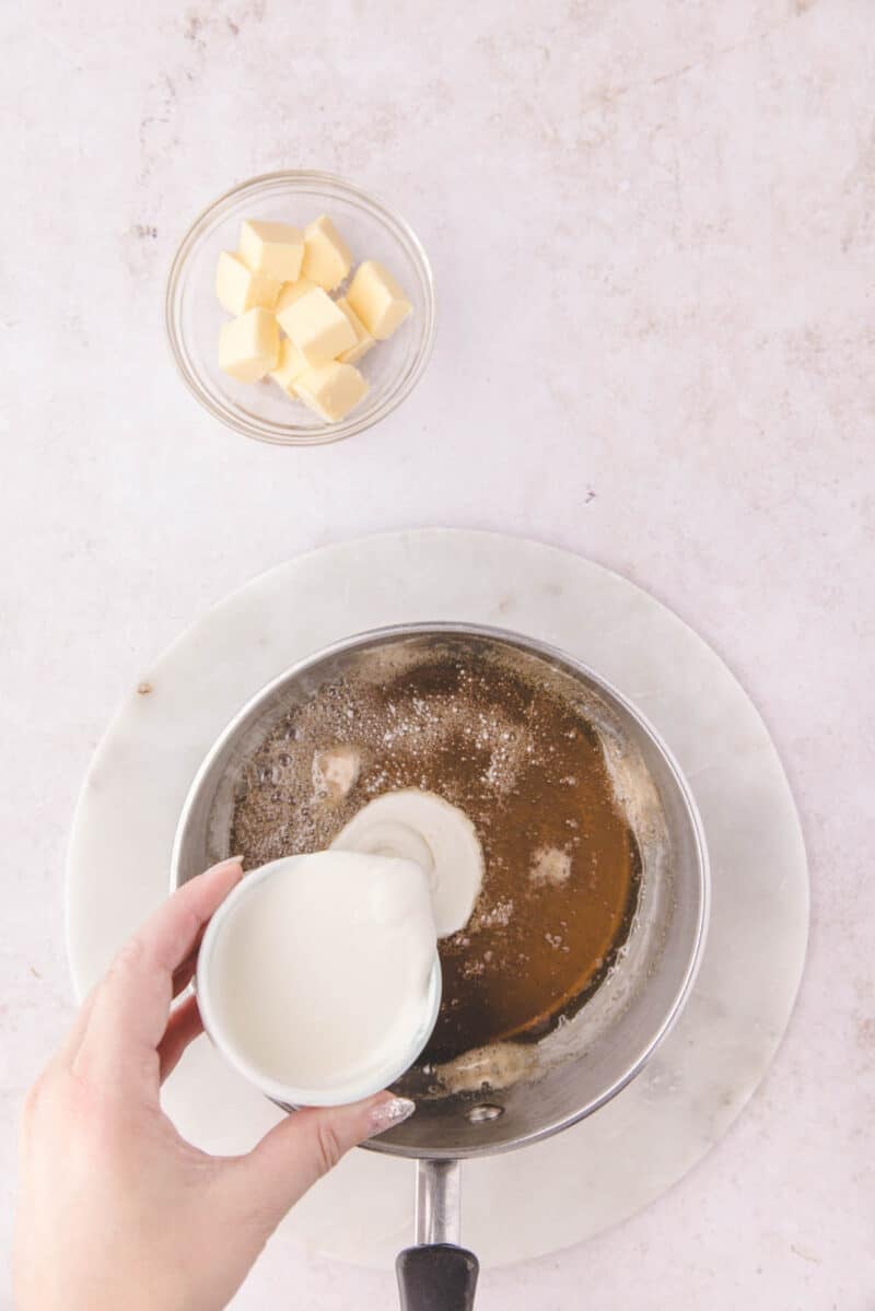A hand pours cream into a saucepan containing browned butter, with a small bowl of butter cubes nearby.