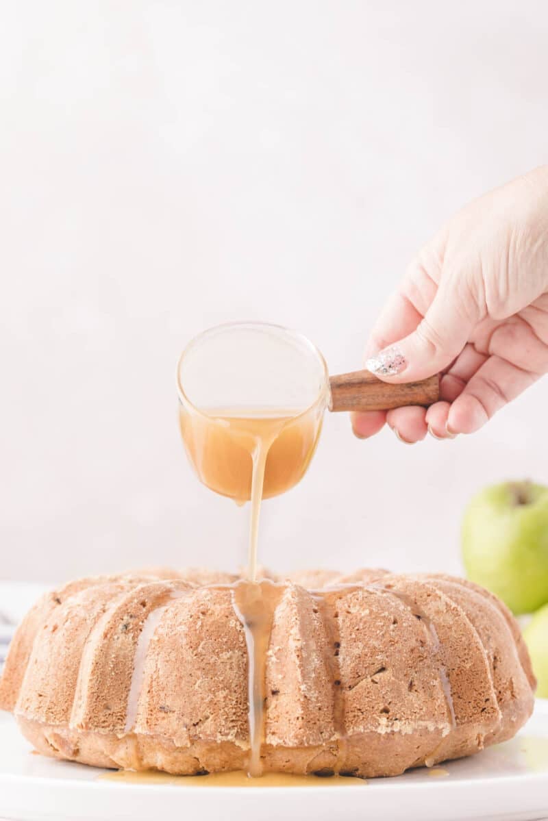Hand pouring caramel glaze from a glass pitcher onto a bundt cake.