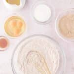 An overhead view of baking ingredients, including flour in a large bowl with a whisk, eggs, sugar, oil, and vanilla extract.