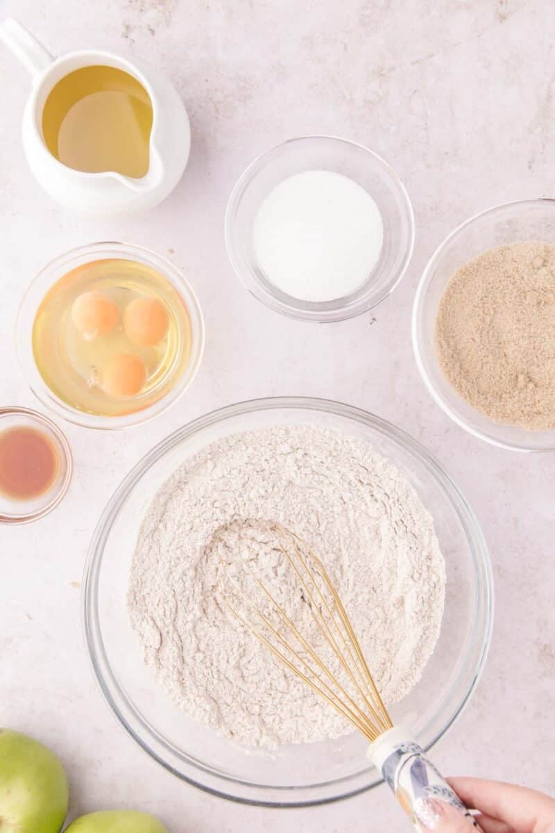An overhead view of baking ingredients, including flour in a large bowl with a whisk, eggs, sugar, oil, and vanilla extract.