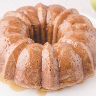 A glazed ring bundt cake on a white plate with a green apple in the background.