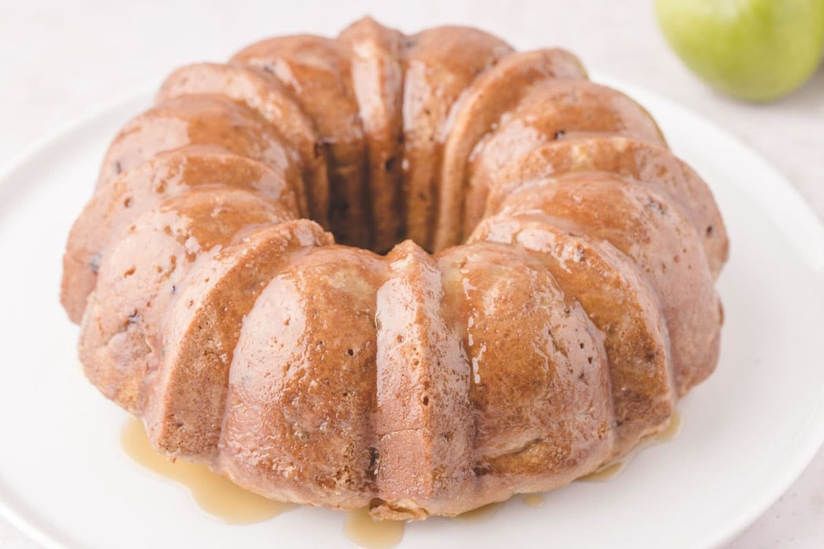 A glazed ring bundt cake on a white plate with a green apple in the background.