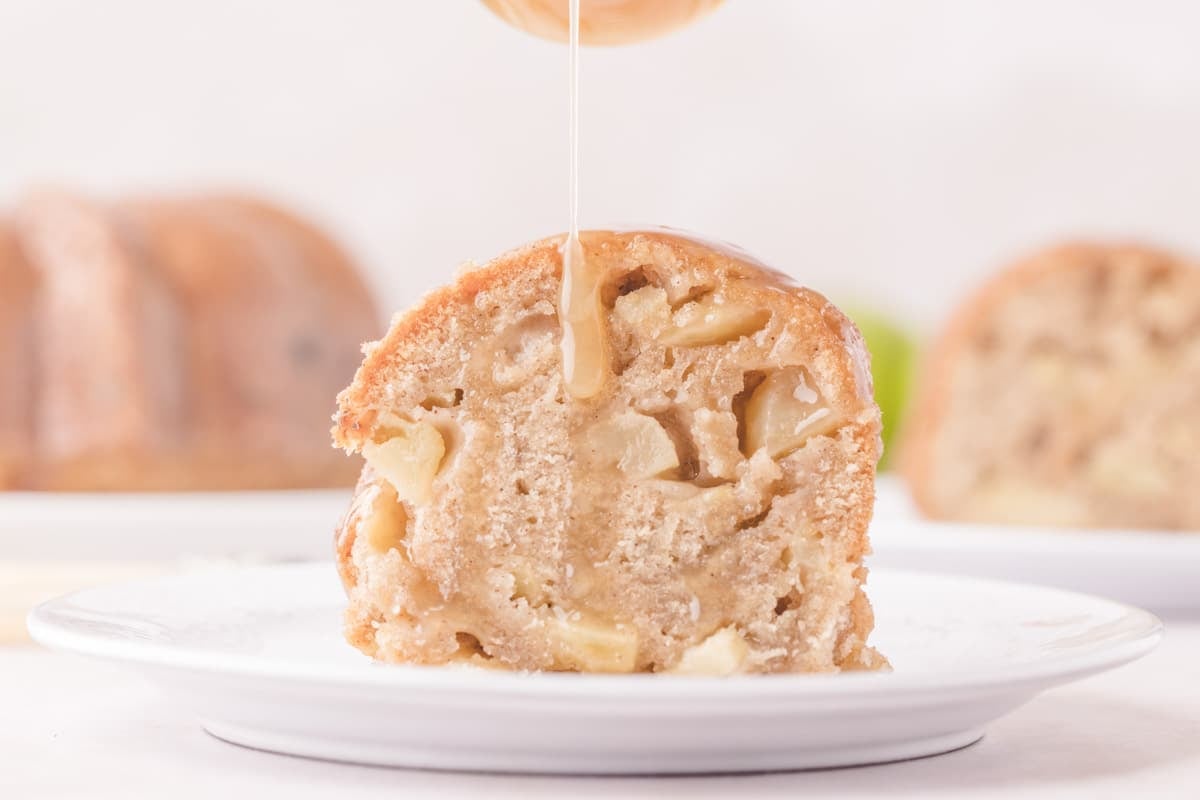 A slice of apple bundt cake on a white plate being drizzled with syrup.