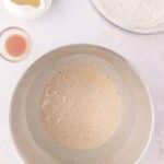 A mixing bowl containing a foamy liquid, surrounded by a pitcher of oil, a small dish of extract, and a bowl of flour.