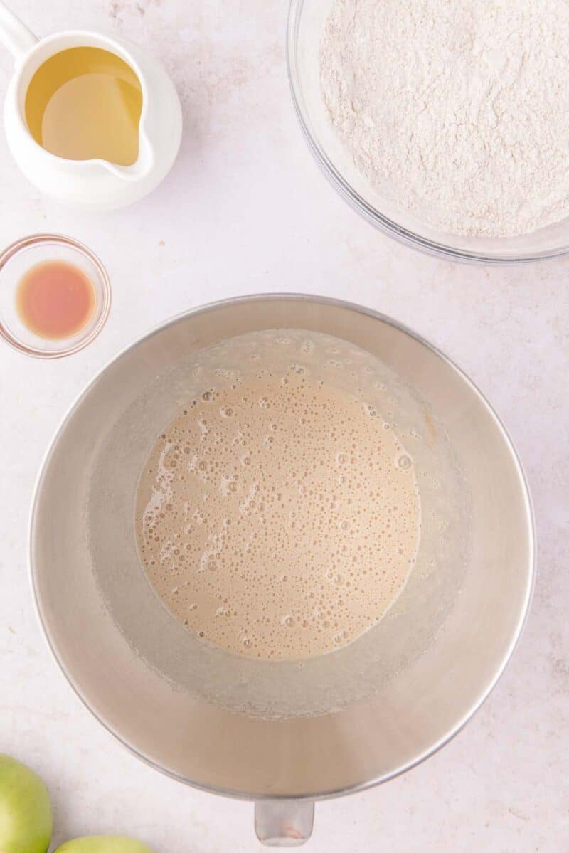 A mixing bowl containing a foamy liquid, surrounded by a pitcher of oil, a small dish of extract, and a bowl of flour.
