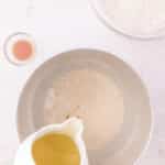 Oil being poured from a small pitcher into a metal mixing bowl containing a tan, bubbly liquid, with a bowl of flour and a small dish of vanilla nearby.