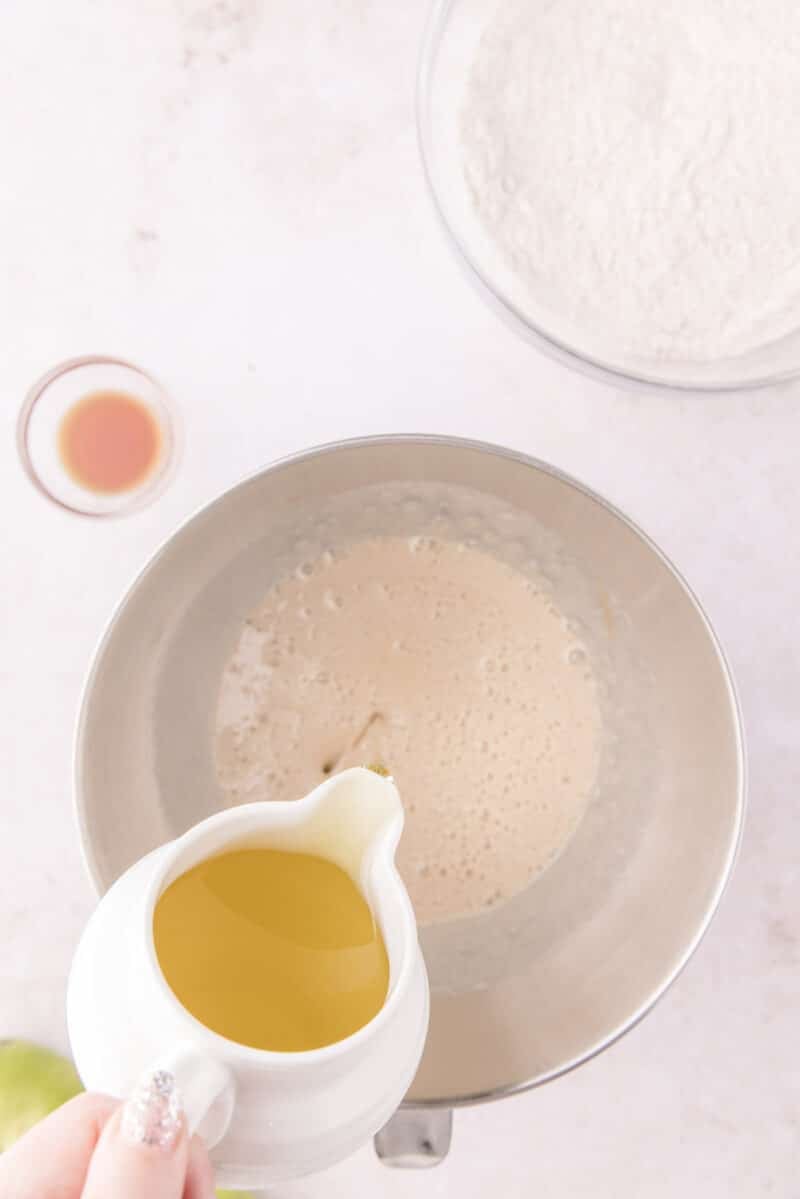 Oil being poured from a small pitcher into a metal mixing bowl containing a tan, bubbly liquid, with a bowl of flour and a small dish of vanilla nearby.