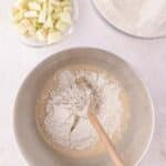 A metal mixing bowl containing wet batter and flour being stirred with a wooden spoon, with separate bowls of chopped apples and flour nearby.