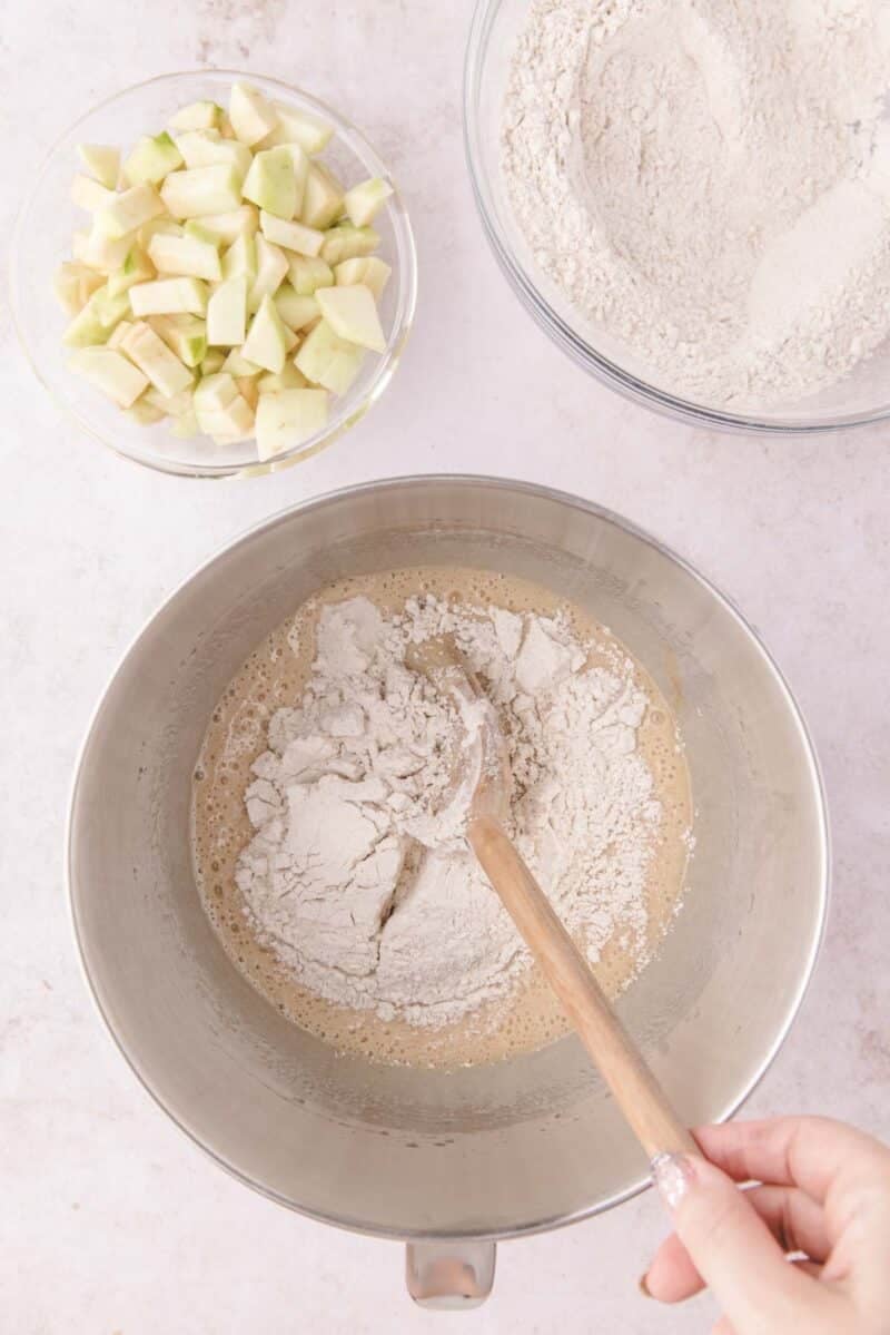 A metal mixing bowl containing wet batter and flour being stirred with a wooden spoon, with separate bowls of chopped apples and flour nearby.