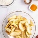 A large glass bowl filled with sliced apples tossed in cinnamon, with smaller bowls containing flour, sugar, and vanilla nearby.