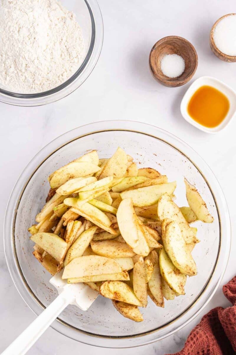 A large glass bowl filled with sliced apples tossed in cinnamon, with smaller bowls containing flour, sugar, and vanilla nearby.