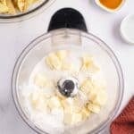 A food processor bowl containing flour and cubes of butter, with a bowl of sliced apples and small dishes of liquid nearby.