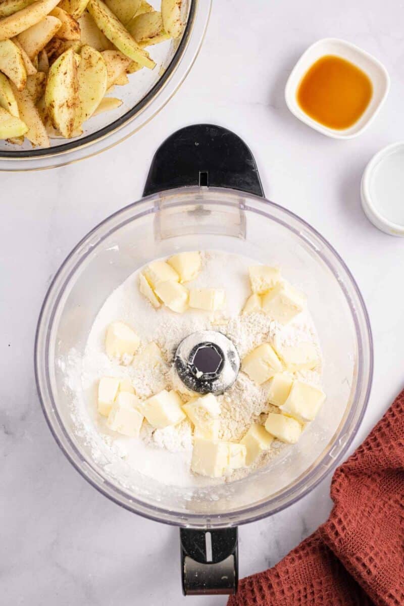 A food processor bowl containing flour and cubes of butter, with a bowl of sliced apples and small dishes of liquid nearby.