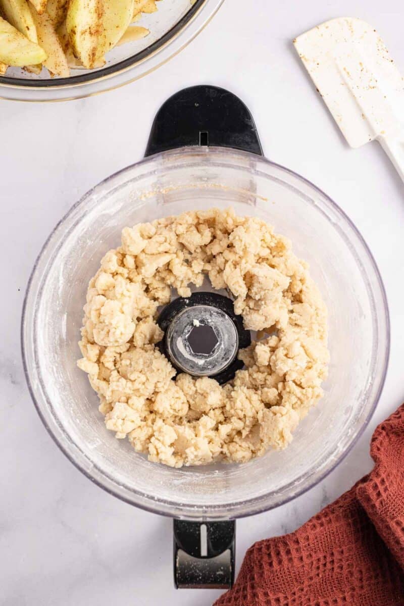 Crumbled pie dough inside a food processor bowl, with sliced apples and a spatula nearby.